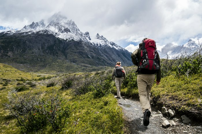 Two hikers with backpacks walking a trail towards snow-capped mountains