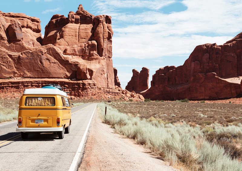A yellow camper van driving down an open road between red rock formations